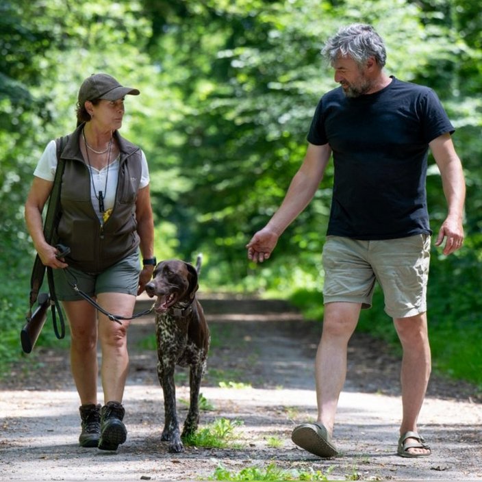 Ein Hund läuft an der Leine einer Frau, während ein Mann neben ihnen geht. Sie befinden sich auf einem Waldweg, umgeben von grünen Bäumen.