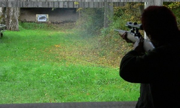 Eine Person zielt mit einem Gewehr auf eine entfernte Zielscheibe. Der Schießstand ist von Bäumen und Gras umgeben, die Umgebung wirkt herbstlich und bewaldet.