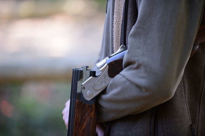 Eine Flinte liegt geöffnet im Arm einer Person, die eine dunkelgrüne Jacke trägt. Die Umgebung ist verschwommen und sieht nach Natur aus.