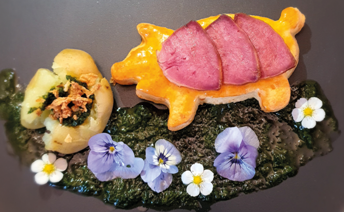 Gebäck in Schweineform, belegt mit rosa Fleischscheiben, ruht auf grünem Pesto. Daneben eine gefüllte Kartoffel. Umgeben von violetten und weißen Blüten auf einem dunklen Teller.