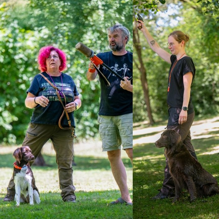 Personen mit Hunden trainieren im Freien mit Trainingsgerät. Links steht eine Person mit rotem Haar neben einem Hund, rechts hält eine Person mit Brille ein Gerät hoch, Hund sitzt davor.