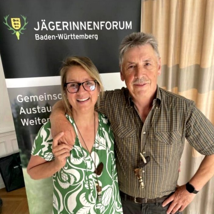 Ein Mann und eine Frau stehen lächelnd nebeneinander; er legt den Arm um sie. Im Hintergrund steht ein Banner mit der Aufschrift "Jägerinnenforum Baden-Württemberg, Gemeinschaft, Austausch, Weiterbildung".