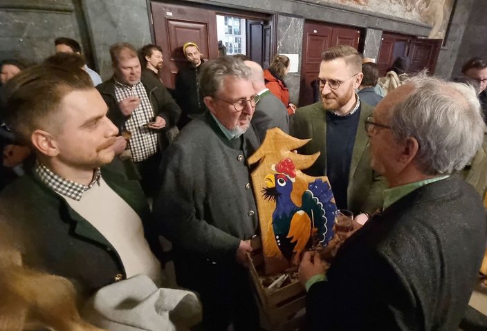 Ein Mann hält ein bunt bemaltes Holzbrett mit einem Huhn, umgeben von mehreren Männern in traditioneller Kleidung, in einem festlichen, mit Menschen gefüllten Raum.