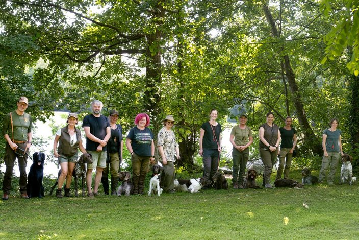 Menschen stehen mit ihren Hunden in einer Reihe, lächelnd und entspannt. Der Hintergrund zeigt üppige grüne Bäume und einen sonnigen Tag in einem Park.