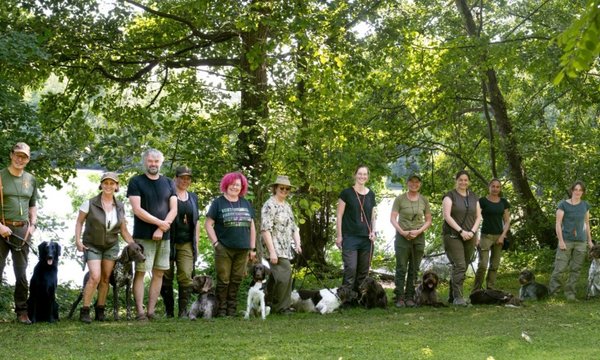 Menschen stehen mit Hunden an der Leine in einer Reihe. Sie lächeln in die Kamera. Im Hintergrund ist ein Wald mit vielen Bäumen zu sehen.