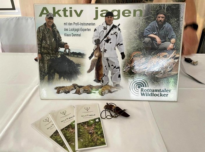 Plakat zeigt Jäger in Tarnkleidung mit Beute. Text: "Aktiv jagen mit den Profi-Instrumenten des Lockjagd-Experten Klaus Demmel", "Rottumtaler Wildlocker". Broschüren "WILD im WALD" liegen daneben.