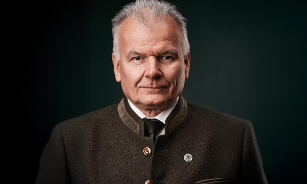 Ein älterer Mann trägt einen grauen Trachtenanzug und schaut ruhig in die Kamera. Er befindet sich vor einem dunklen, einfarbigen Hintergrund.